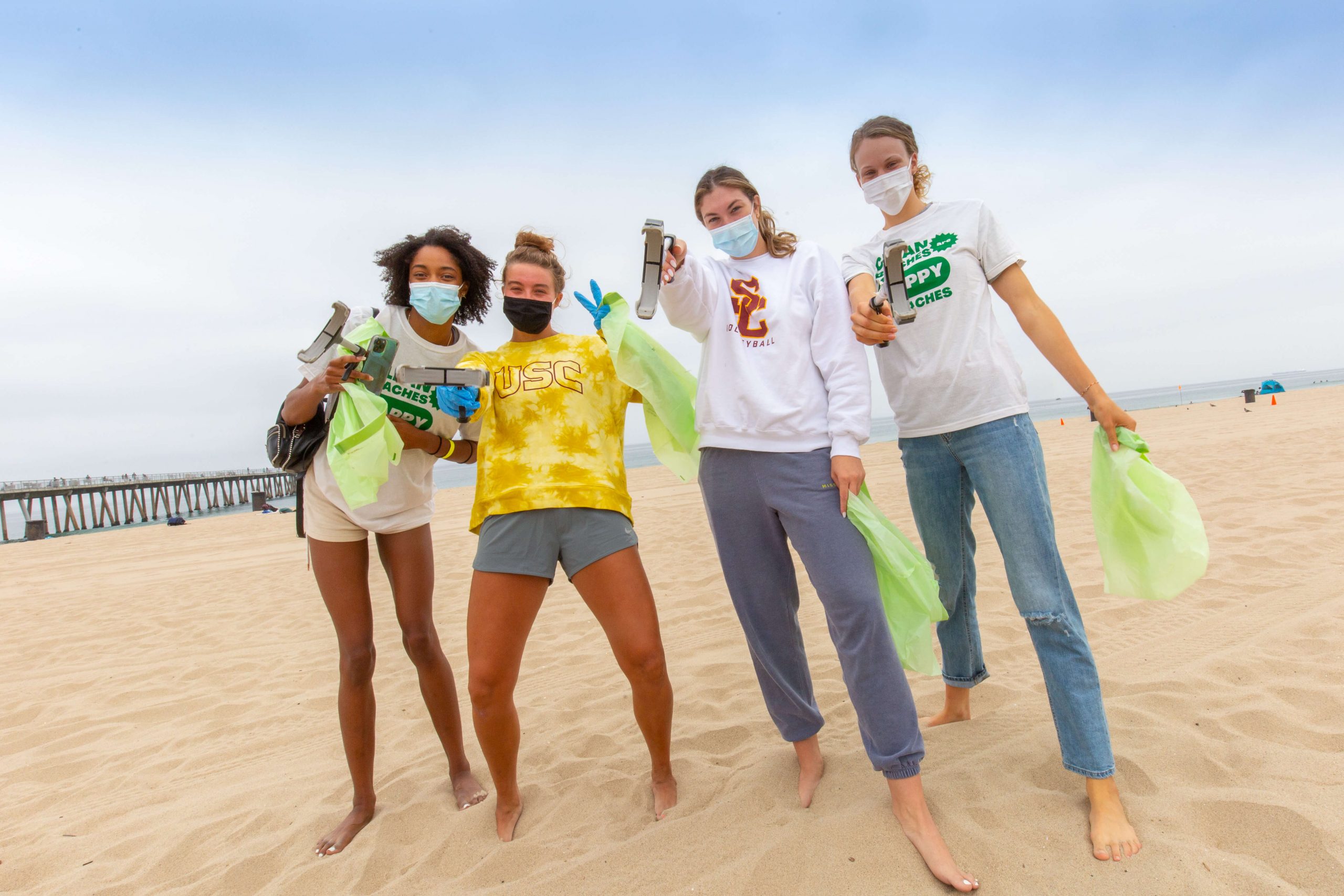 a group of students holding green trash bags and trash scoopers poses together on a beach