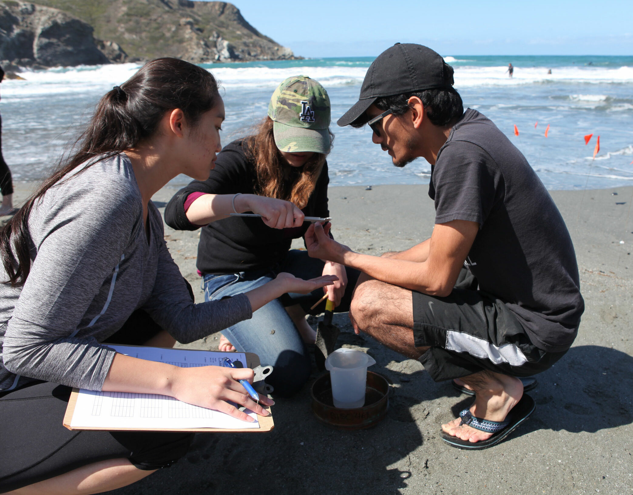 Three students wearing casual clothing crouch on the beach as they take soil samples.