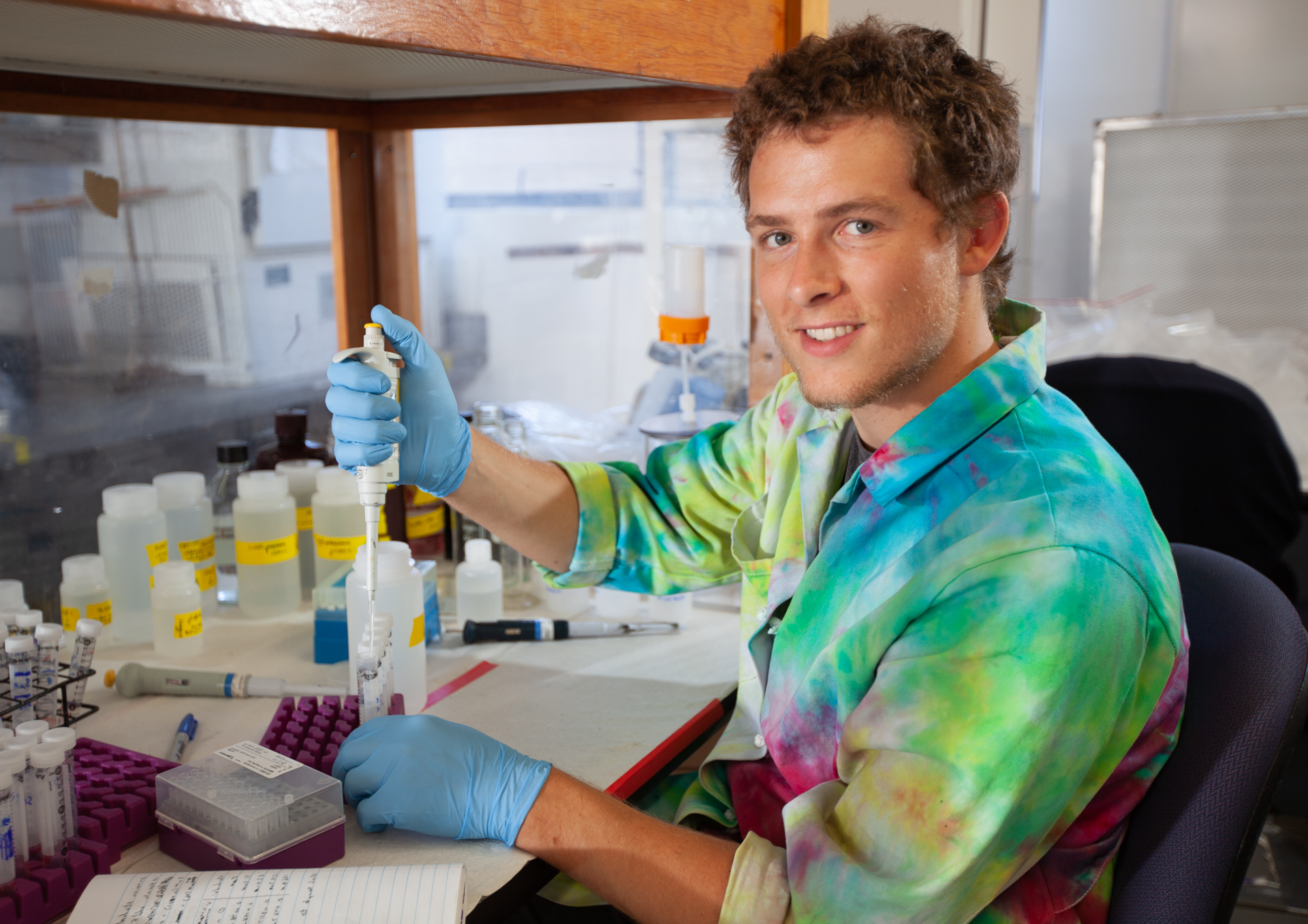 A student wearing a tie-dyed lab coat uses a pipette to fill test tubes with samples