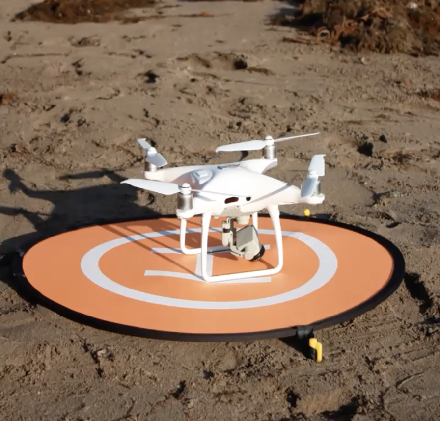 A small white drone sits on a circular landing pad on the sand