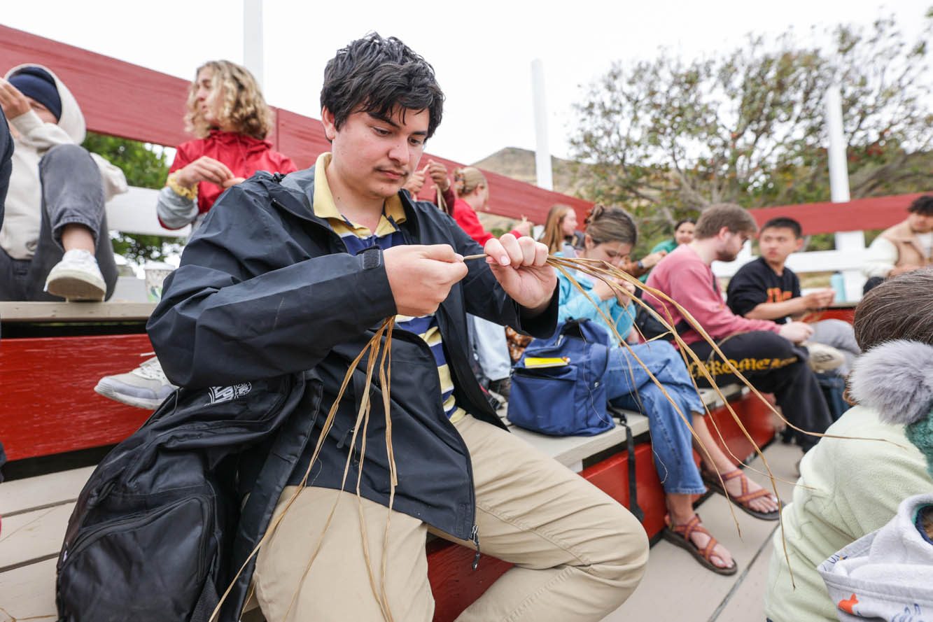 a student wearing a black windbreaker and tan pants sits on a red bench wile separating and braiding plant fibers