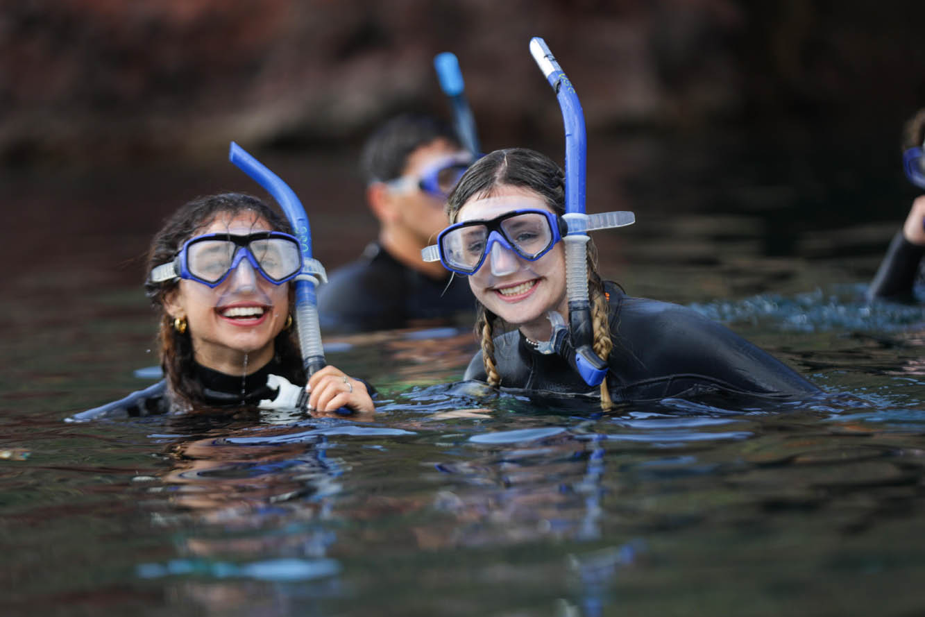two students wearing snorkels and wetsuits smile as they poke their heads above the water