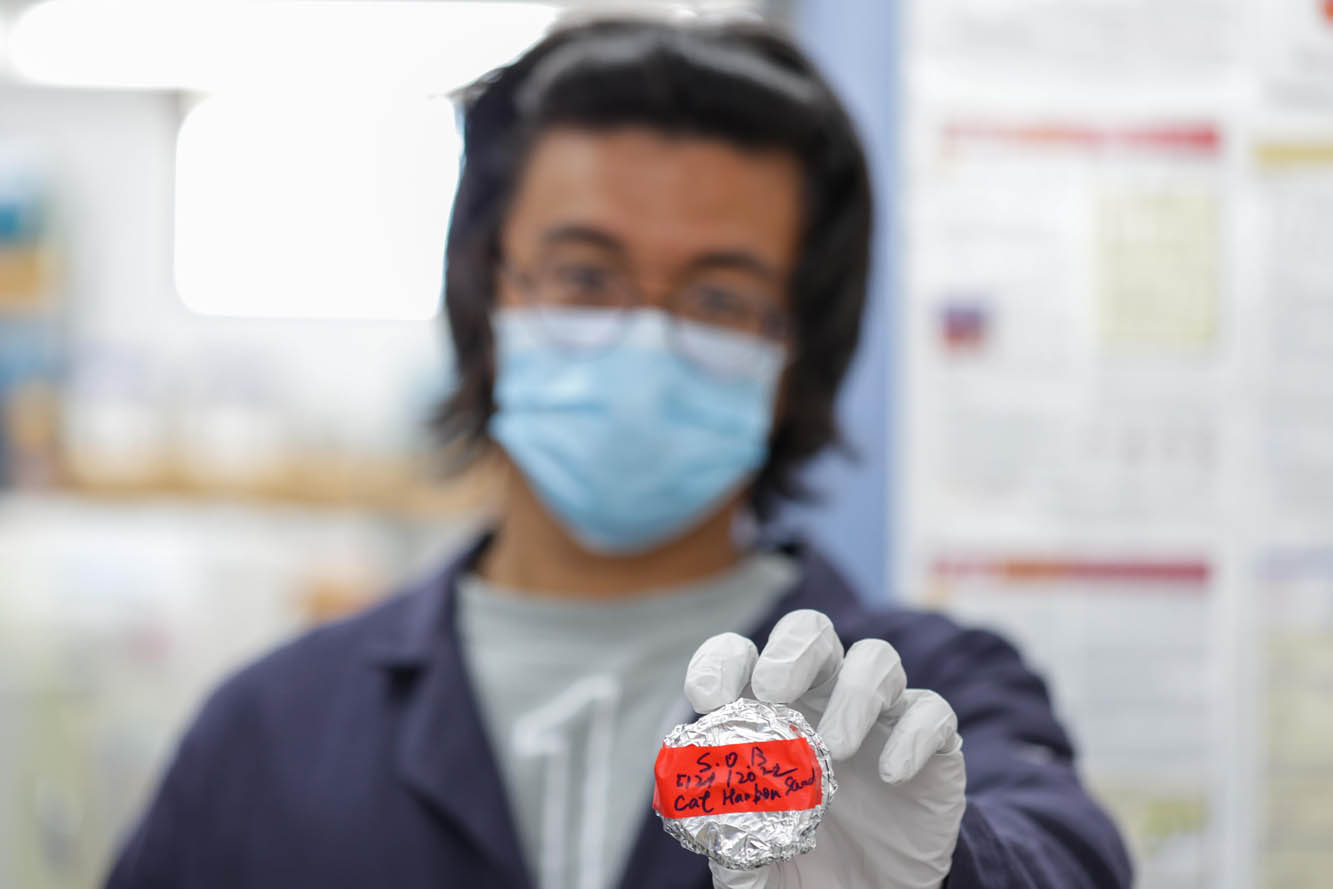 a student wearing a mask and blue lab coat holds out a small, foil-covered container with a red label