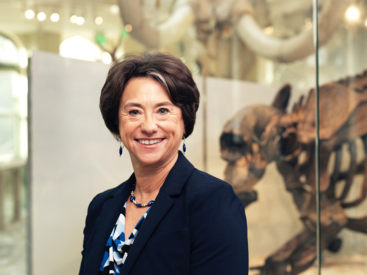 Gale Sinatra, wearing a navy blue blazer and blue-and-white blouse, stands in front of a glass museum case containing the skeleton of a large prehistoric animal