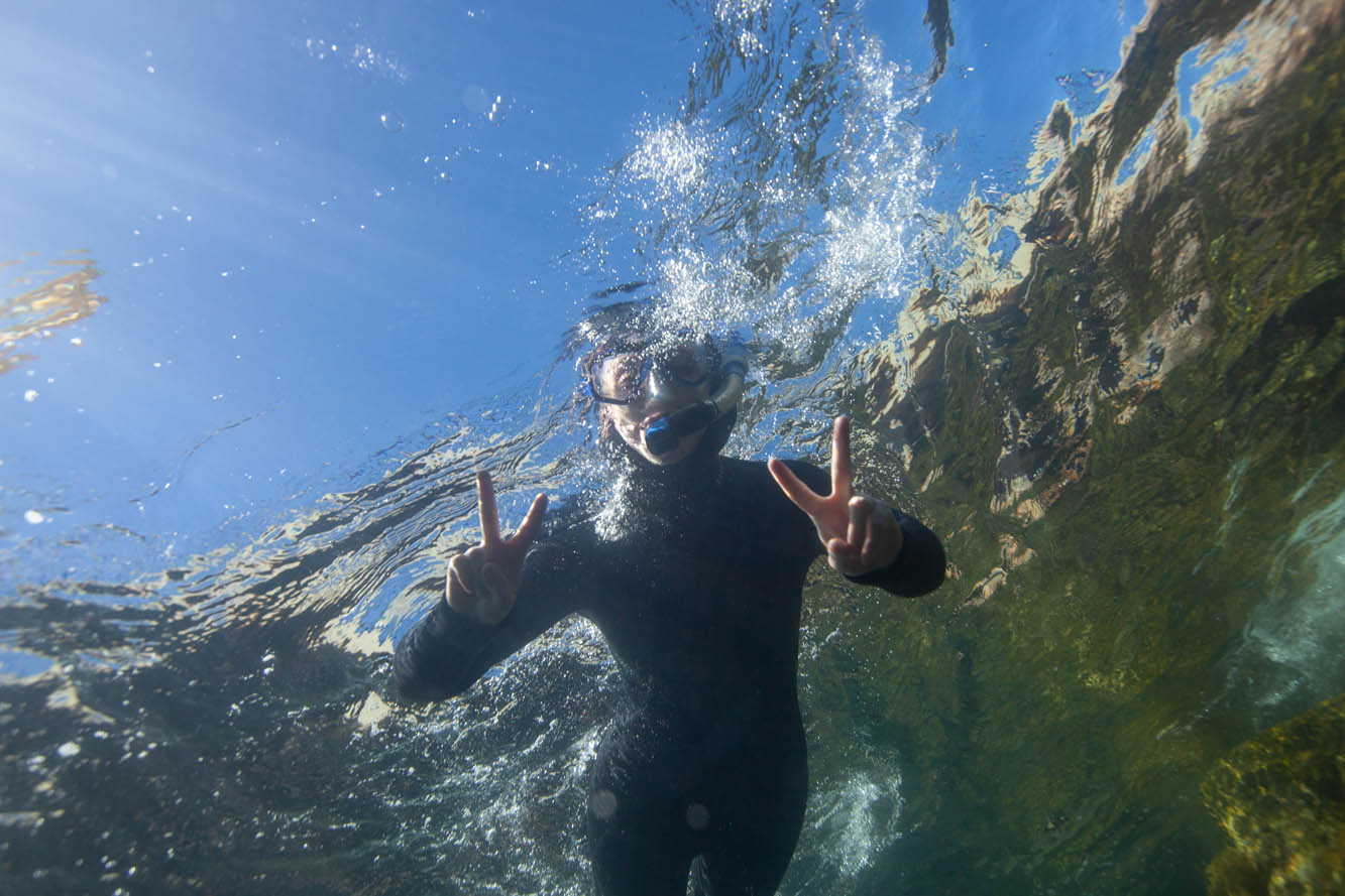 a student floats over the camera in wetsuit and a snorkel, holding up fingers in a victory sign