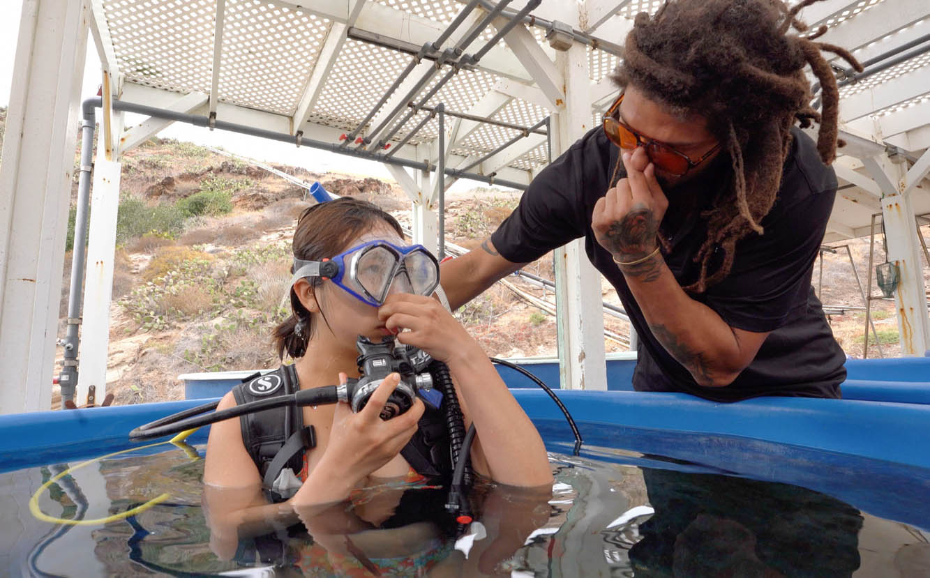 a student wearing dive gear and holding a regulator pinches their nose shut, mimicking the dive instructor standing nearby