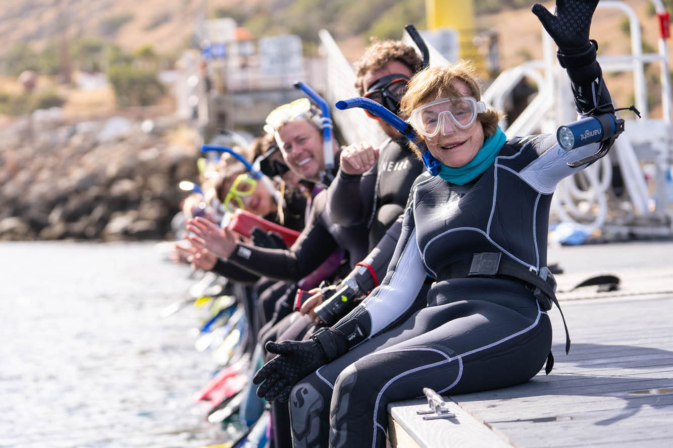 a line of people, wearing wetsuits and snorkels, wave at the camera and smile as they sit on the edge of a dock
