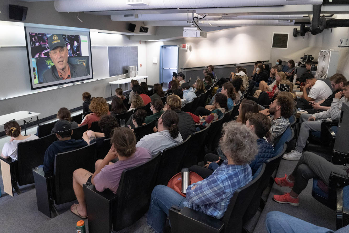 a lecture hall full of students watch a video screen where a man wearing a basball cap and jacket talks to them