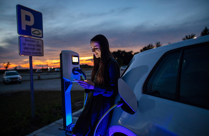 a woman checks her phone at night while waiting for her electric car to charge at a public charging station
