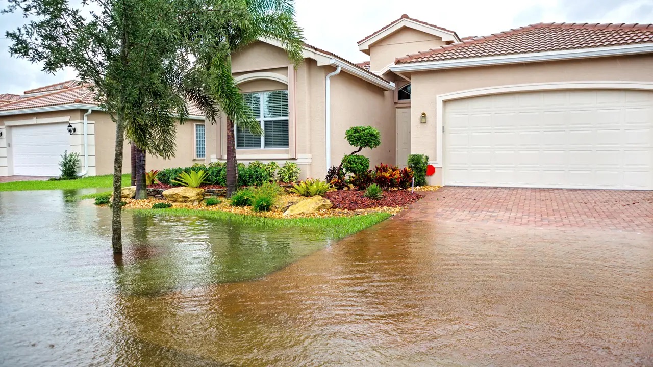 flood waters cover the front lawn and driveway of a tan-colored stucco home with palm trees in the yard