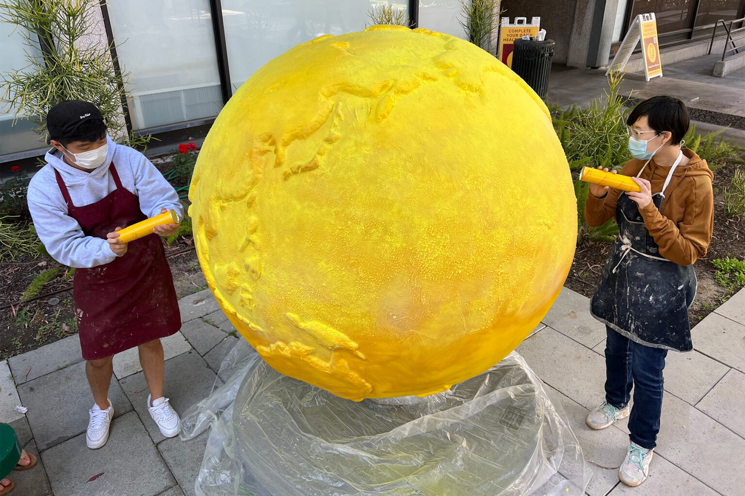 two students wearing masks and aprons stand on either side of a tall, bright-yellow concrete globe as they use large syringes to cover it in bee pollen