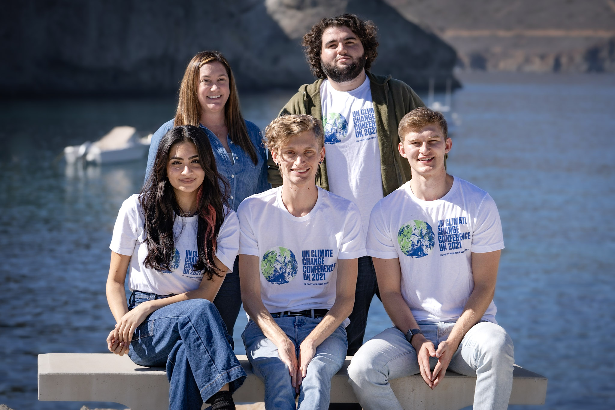 five people, four of them wearing COP26 T-shirts, sit on or stand behind a concrete bench with a cove and hills behind them