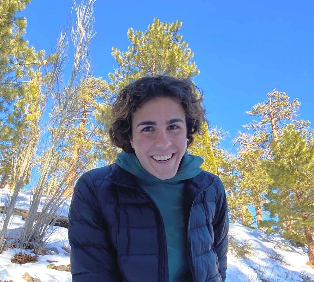 a person smiles while standing in the snow with trees in the background