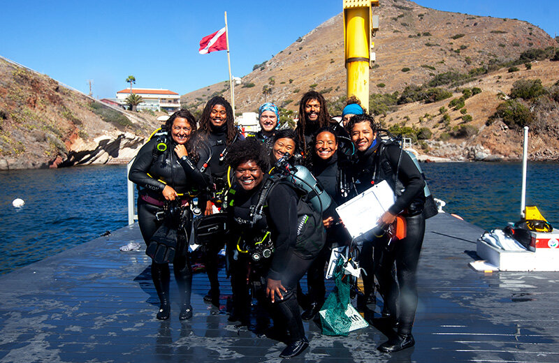 a diverse group of people in dive gear smile as they pose together on a boat dock