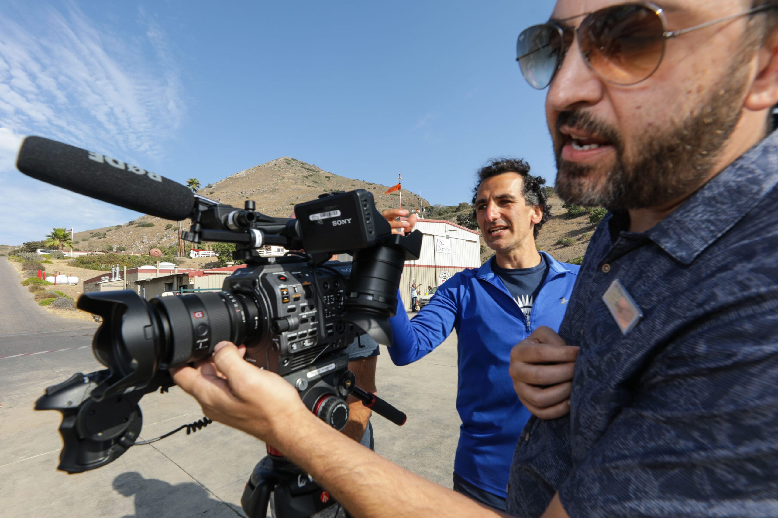 a man in a blue jacket looks through the viewfinder of a video camera while a man wearing sunglasses and a blue shirt adjusts the settings on a lens