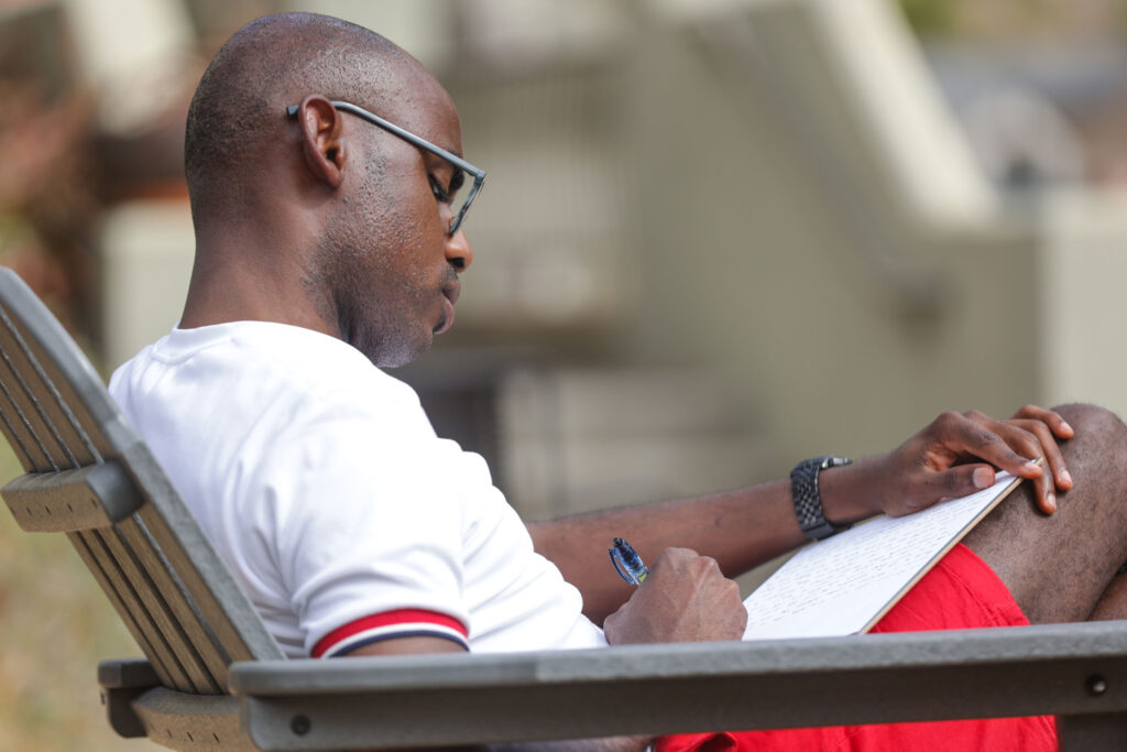 a man wearing glasses, a white shirt, and red shorts writes on a notepad in his lap as he sits on a patio chair outside