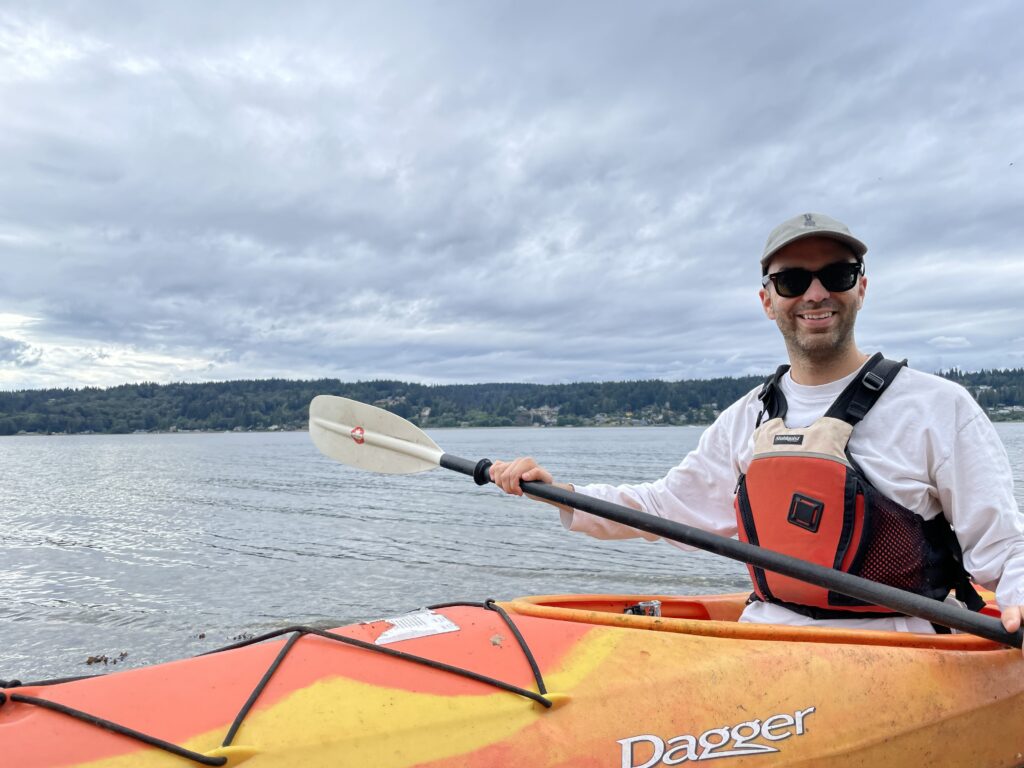 Sean Fraga, wearing a red and black life vest, sits in an orange and yellow kayak with the Pacific Northwest coastline behind him