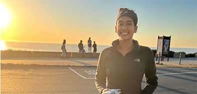 a person in workout clothes smiles while standing in a parking lot with the ocean and sunset behind