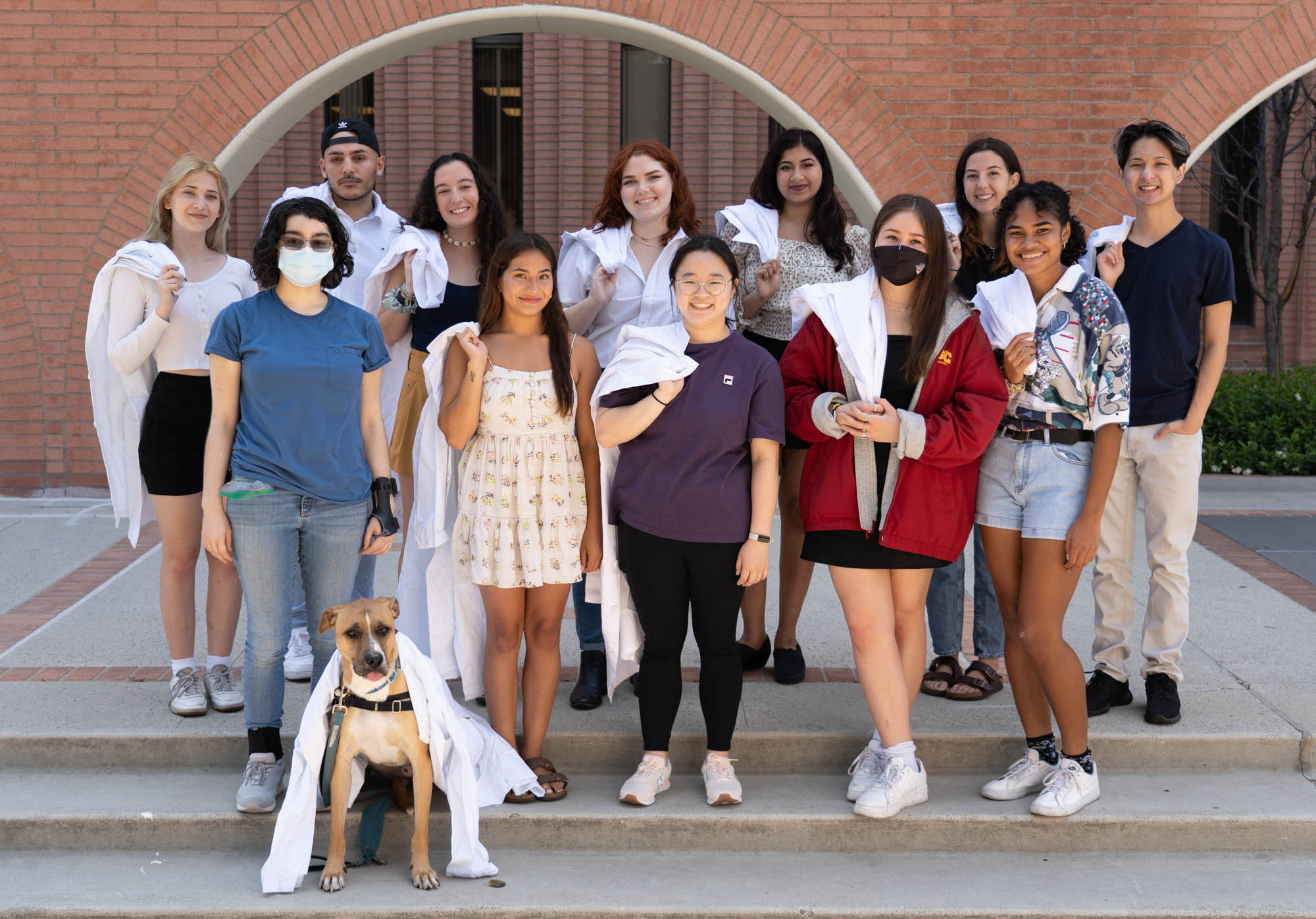 a group of students, some wearing masks or holding white lab coats over their shoulders, stand with a service dog on the concrete steps in front of a red brick building