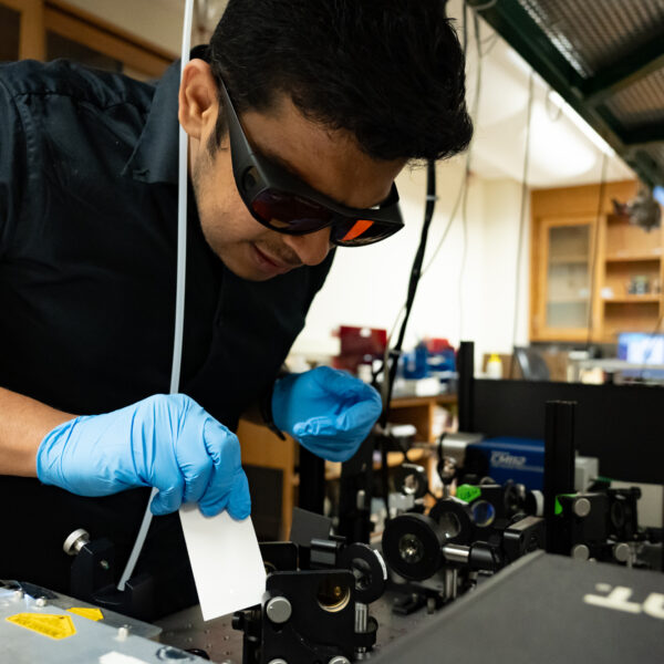 student in blue gloves and protective eye goggles holding a piece of paper in front of a laser