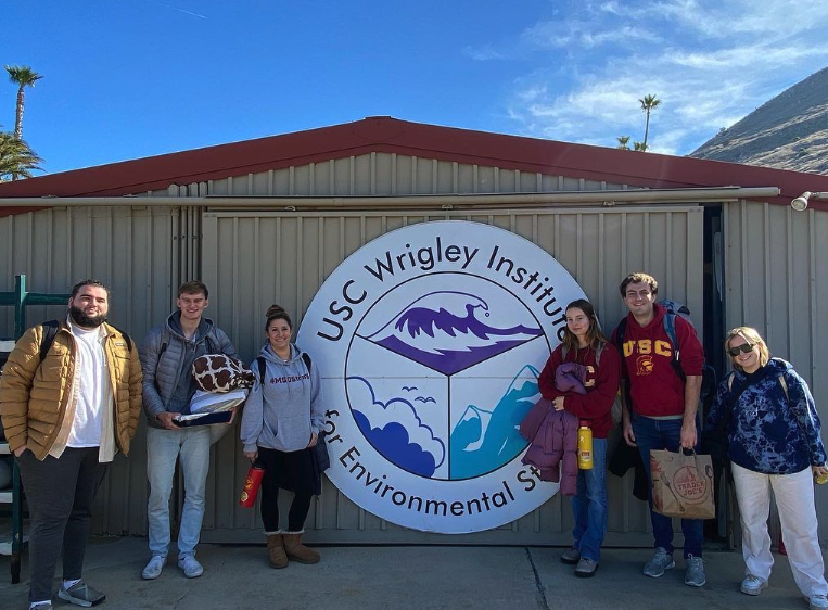 Having just arrived to the Wrigley Marine Science Center on Catalina Island, six climate justice researchers, all wearing backpacks carrying their luggage for the duration of the climate change conference, pose by a large USC Wrigley Institute for Environmental Studies logo.