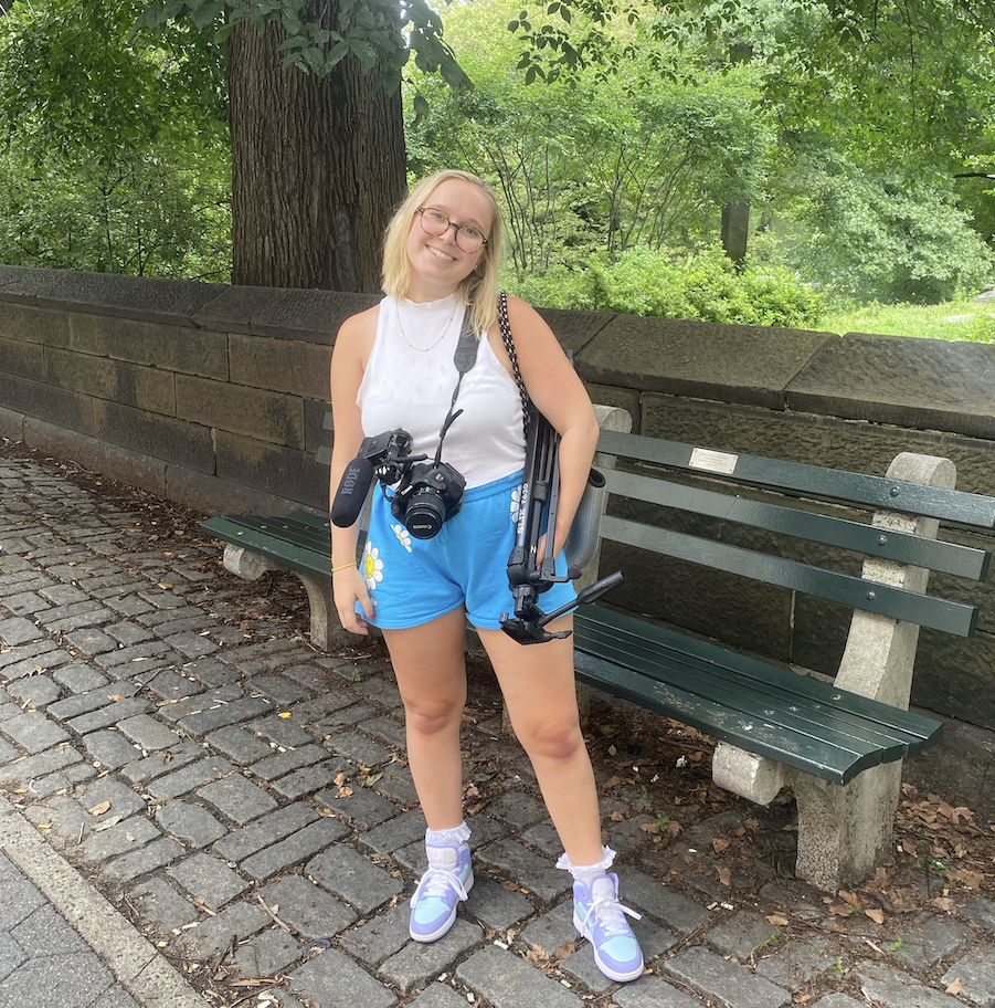 a student wearing a white top and blue shorts stands in Central Park while holding camera equipment