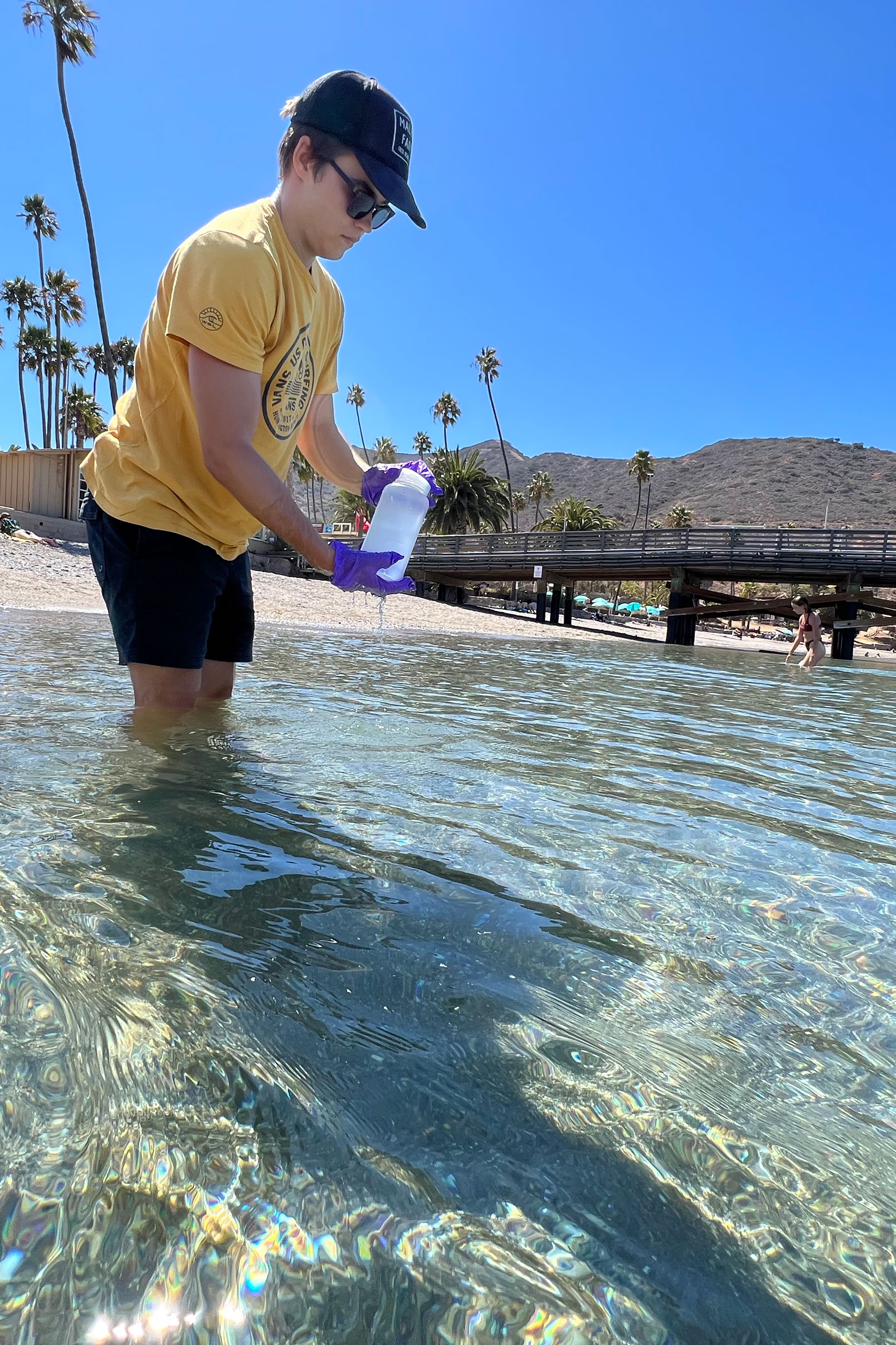 a student holds a sampling bottle while standing in clear, knee-high water with a pier and brown hills in the background