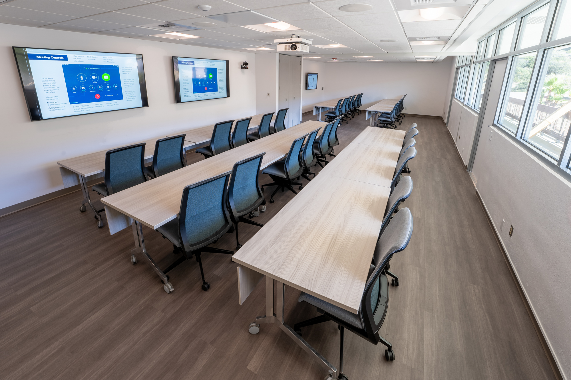 a conference room with large windows, three video monitors, and modular tables and chairs organized into a classroom configuration