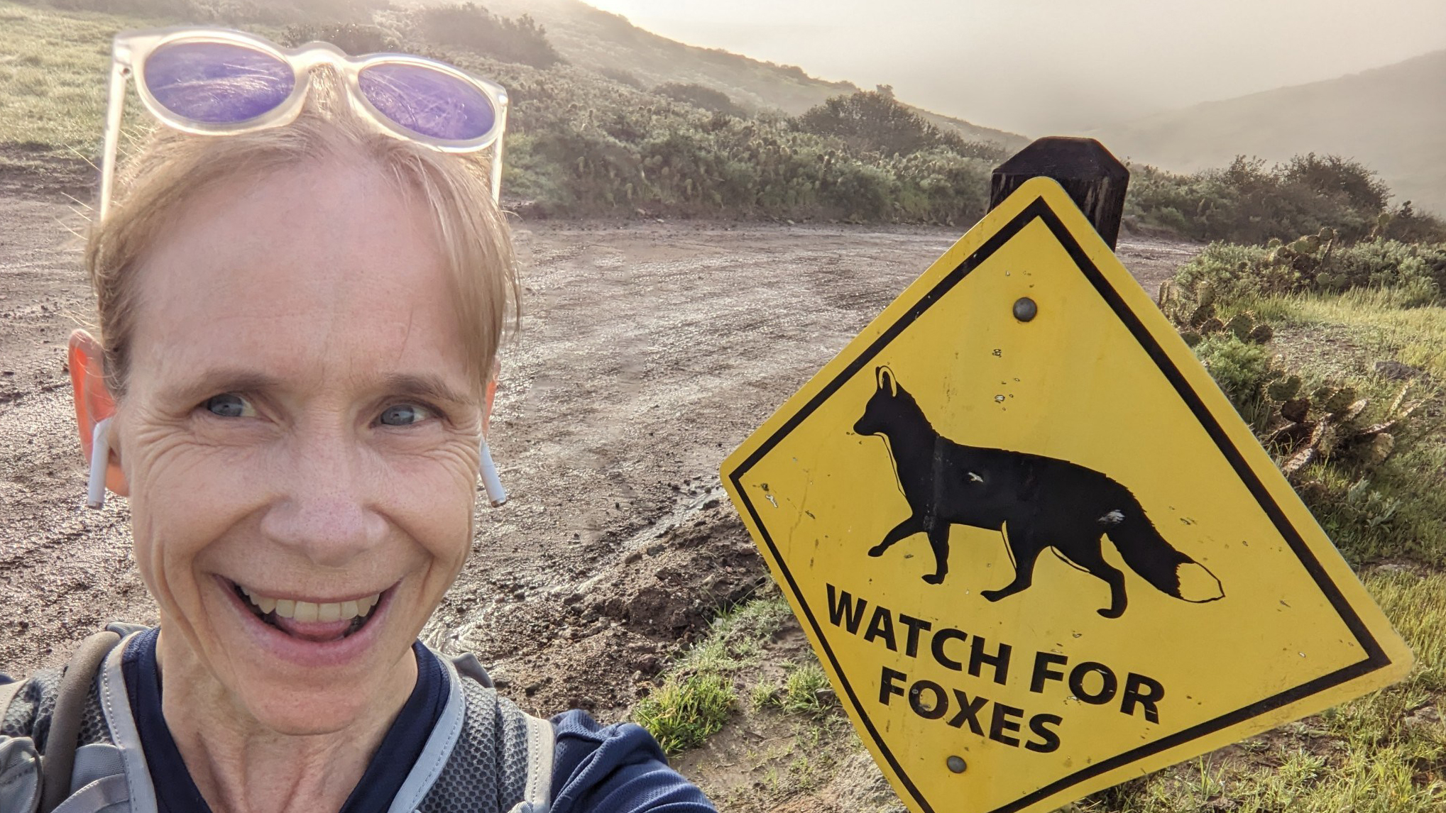 Suzanne Edmands, wearing white sunglasses on her head, smiles while standing in front of a yellow diamond "Watch for Foxes" sign next to a Catalina Island hiking trail