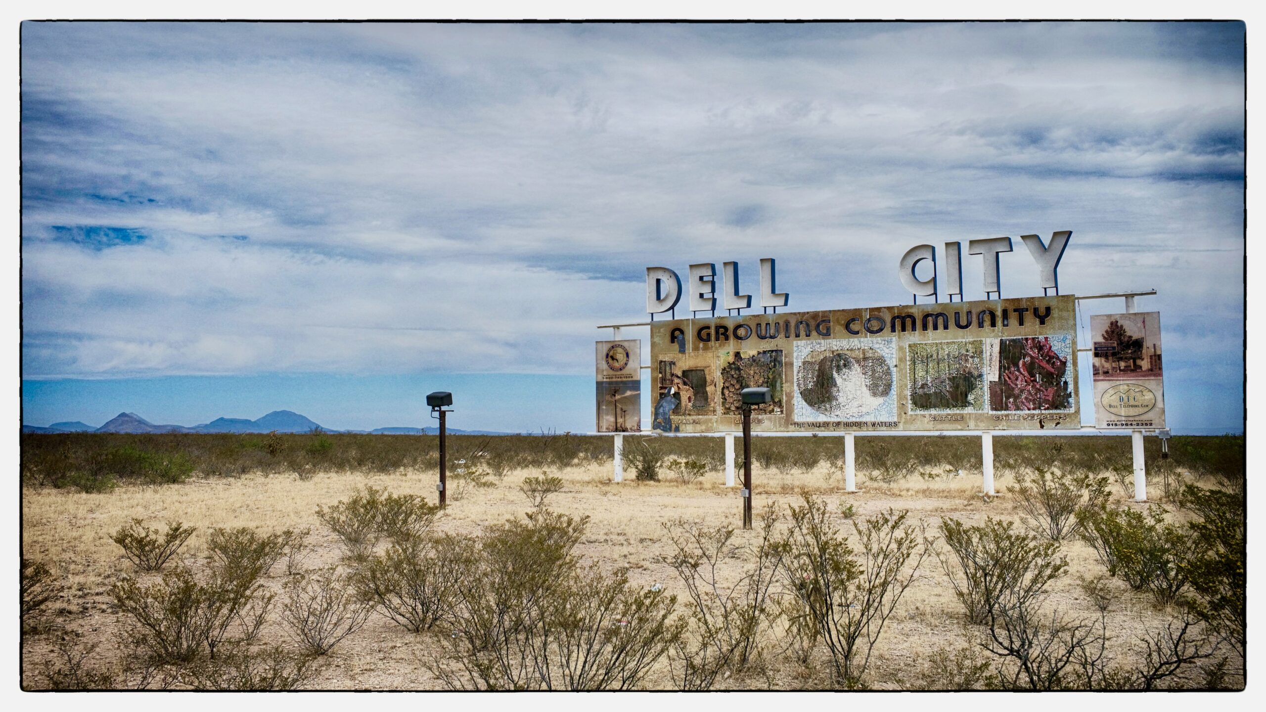 a weathered billboard, topped by the words "Dell City" and displaying faded images of industry and nature, stands in a scrubby desert landscape with mountain peaks and a cloudy blue sky in the background