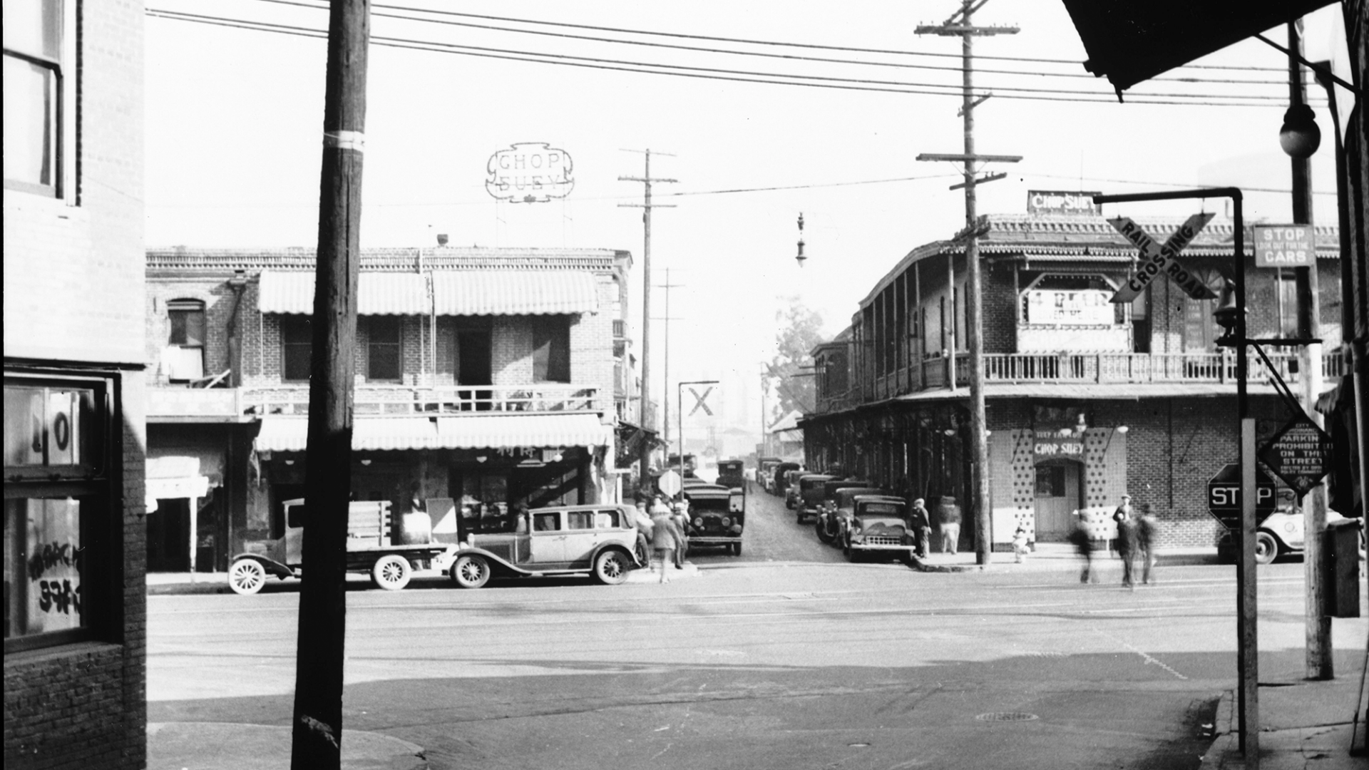 a black-and-white 1935 view of Los Angeles Chinatown, showing people crossing the street and cars parked in front of restaurants and shops
