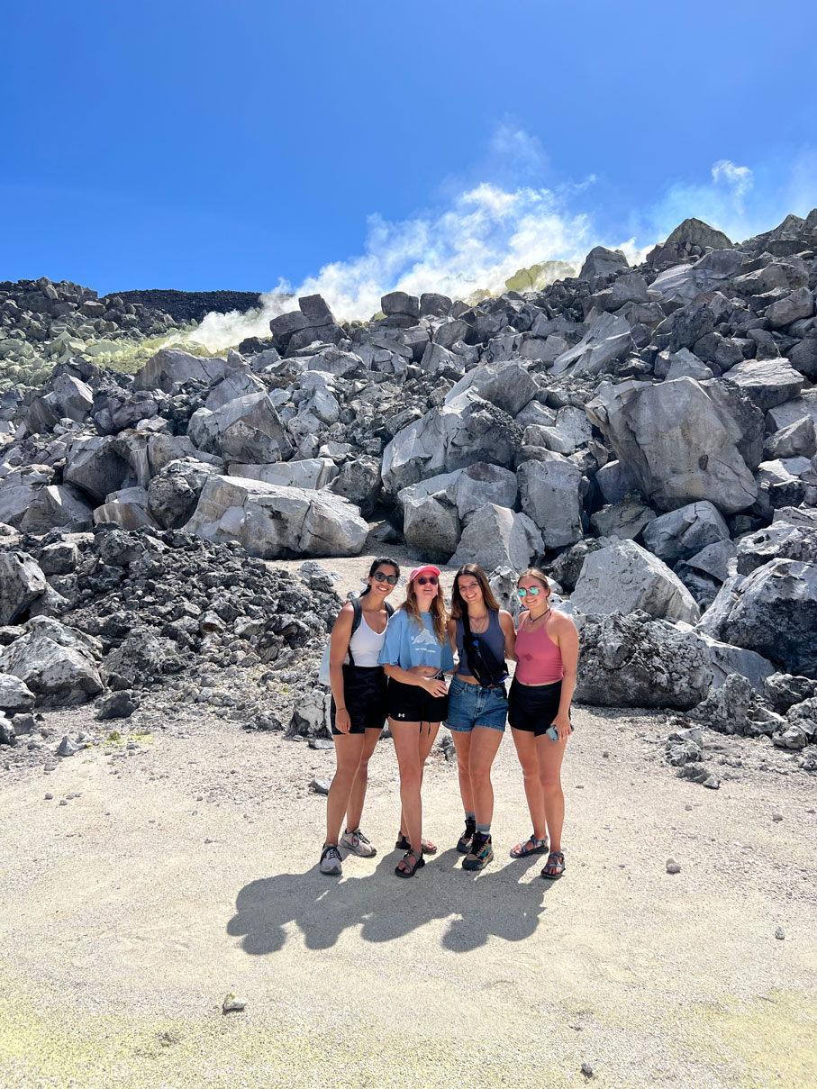 Alisha Soni stands with two friends in front of a gray rockfall with steam rising from the top