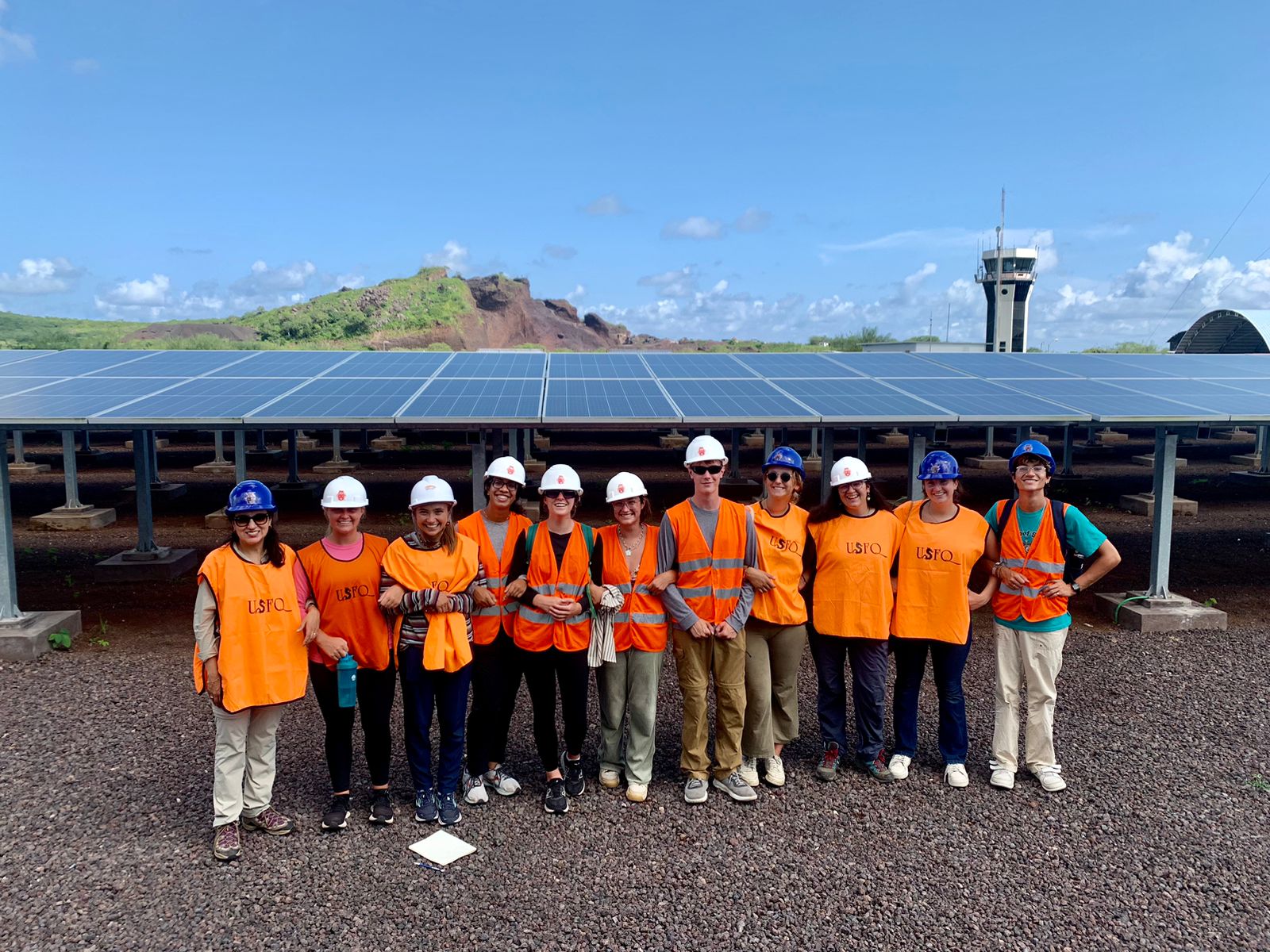 a group of students wearing orange safety vests and hard hats stands in front of a solar panel array