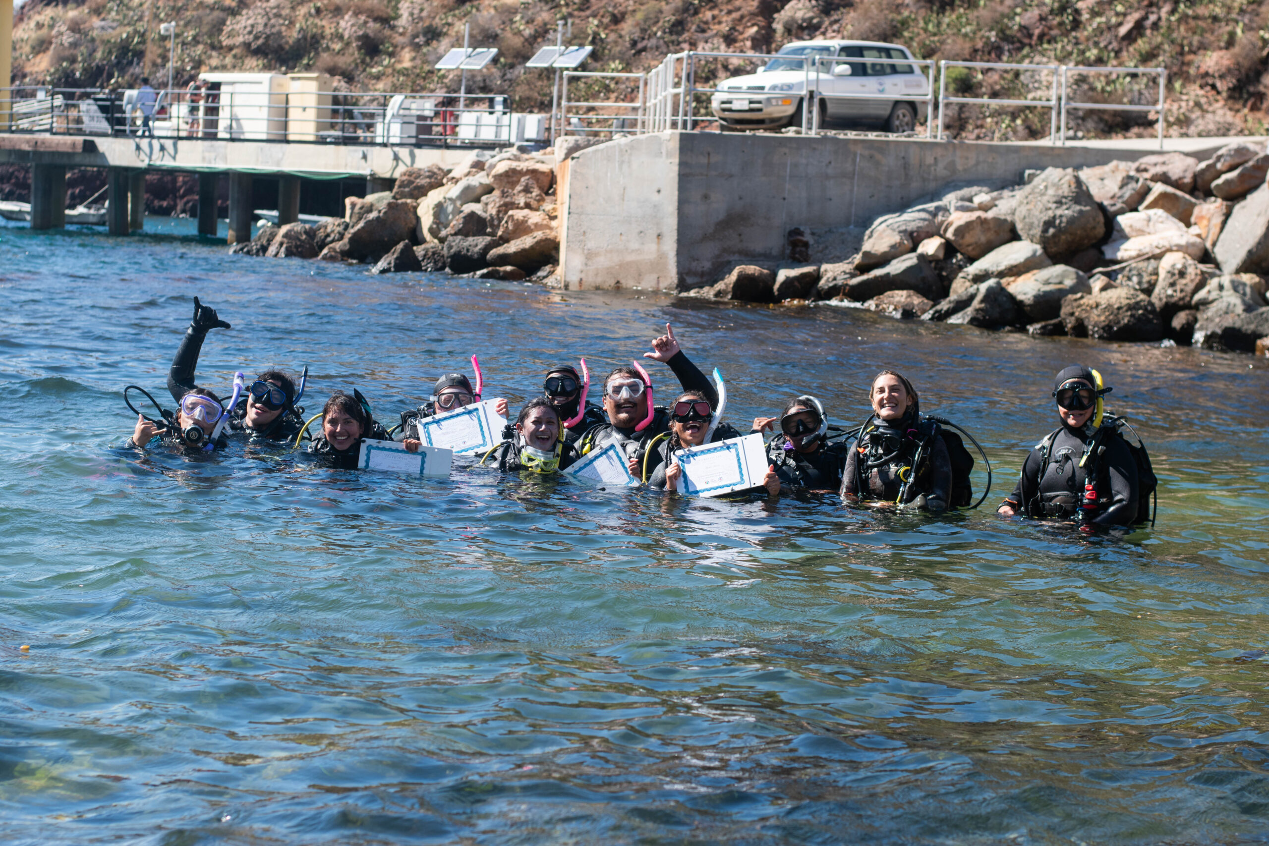 a group of people in dive gear float in a cove as some of the people hold up graduation certificates