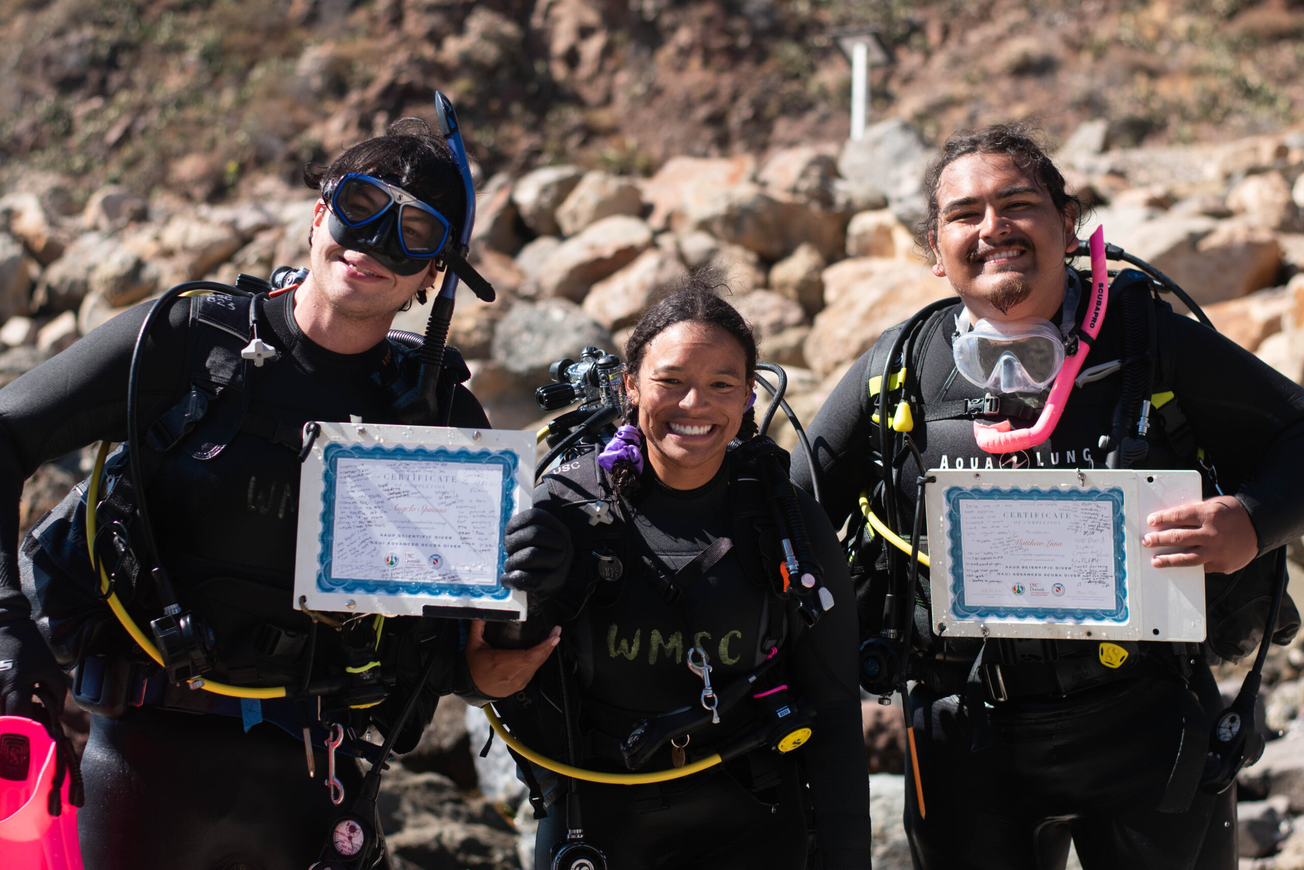 three people in dive gear, two of them holding certificates, stand together by the water