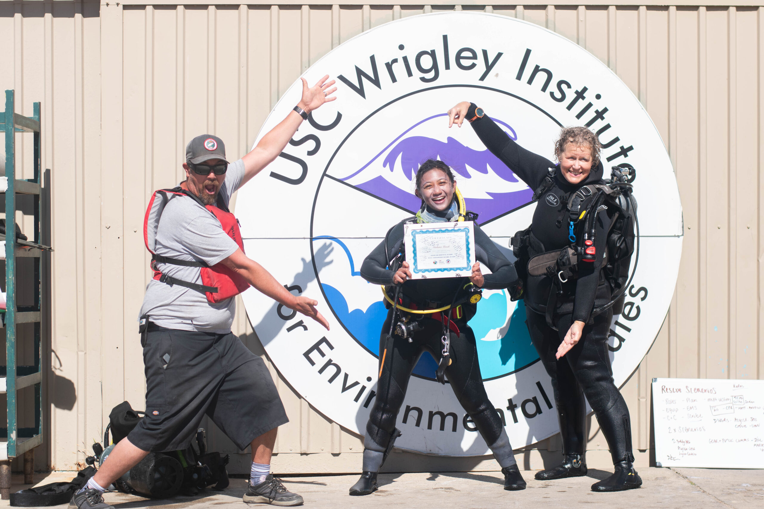a smiling student in dive gear shows off a graduation certificate while standing between two instructors