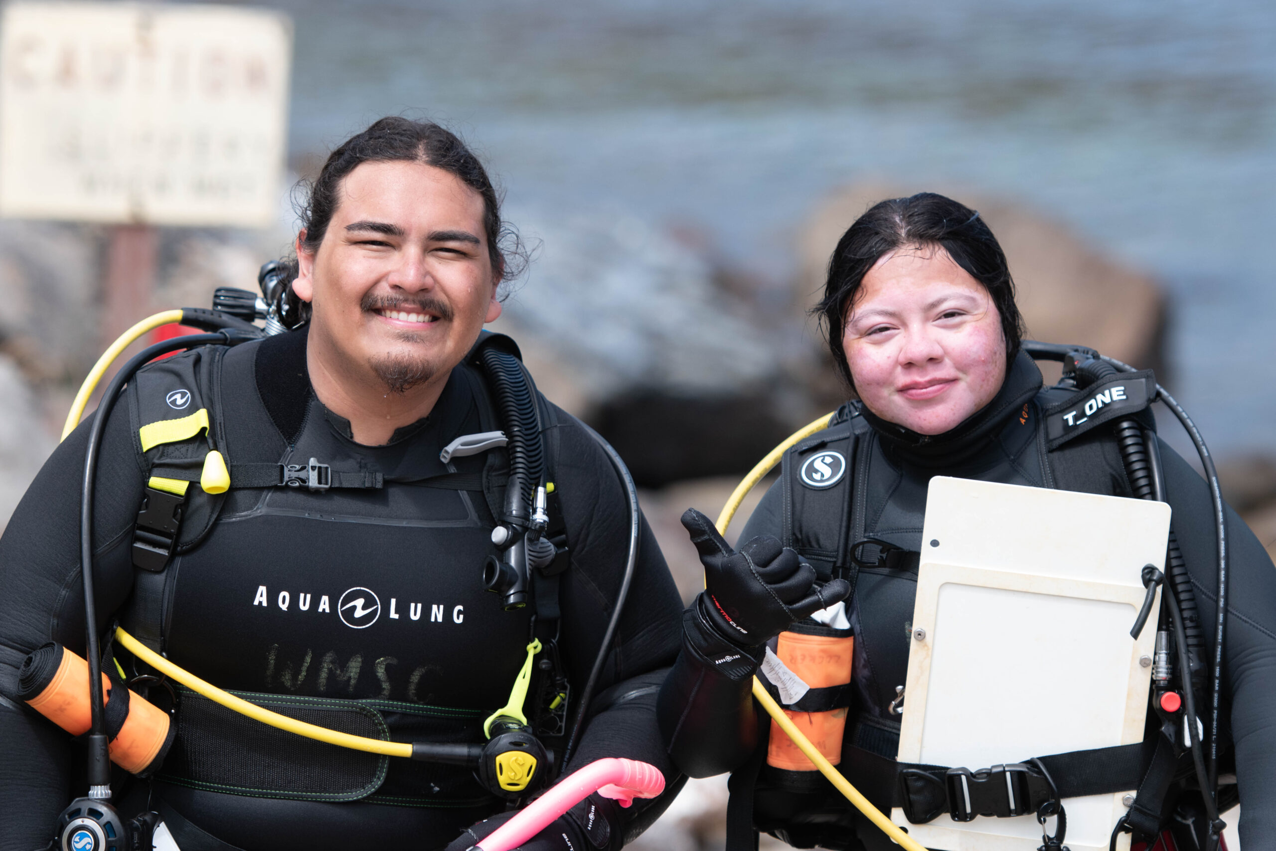 two individuals in dive gear, one holding an underwater whiteboard, stand on a dock