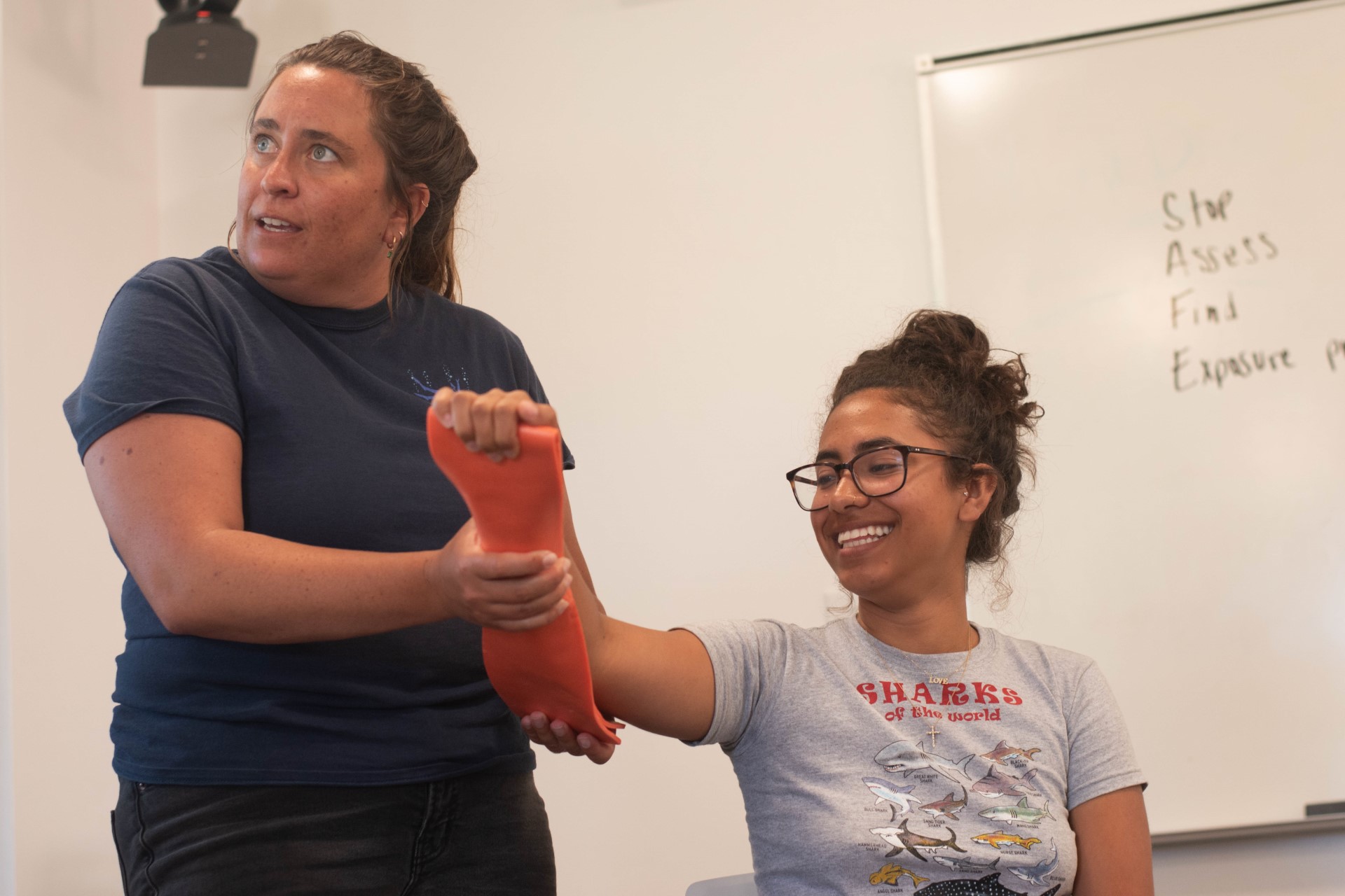 an instructor holds up the arm of a smiling student who is holding onto a bright-orange board as part of a first-aid demonstration