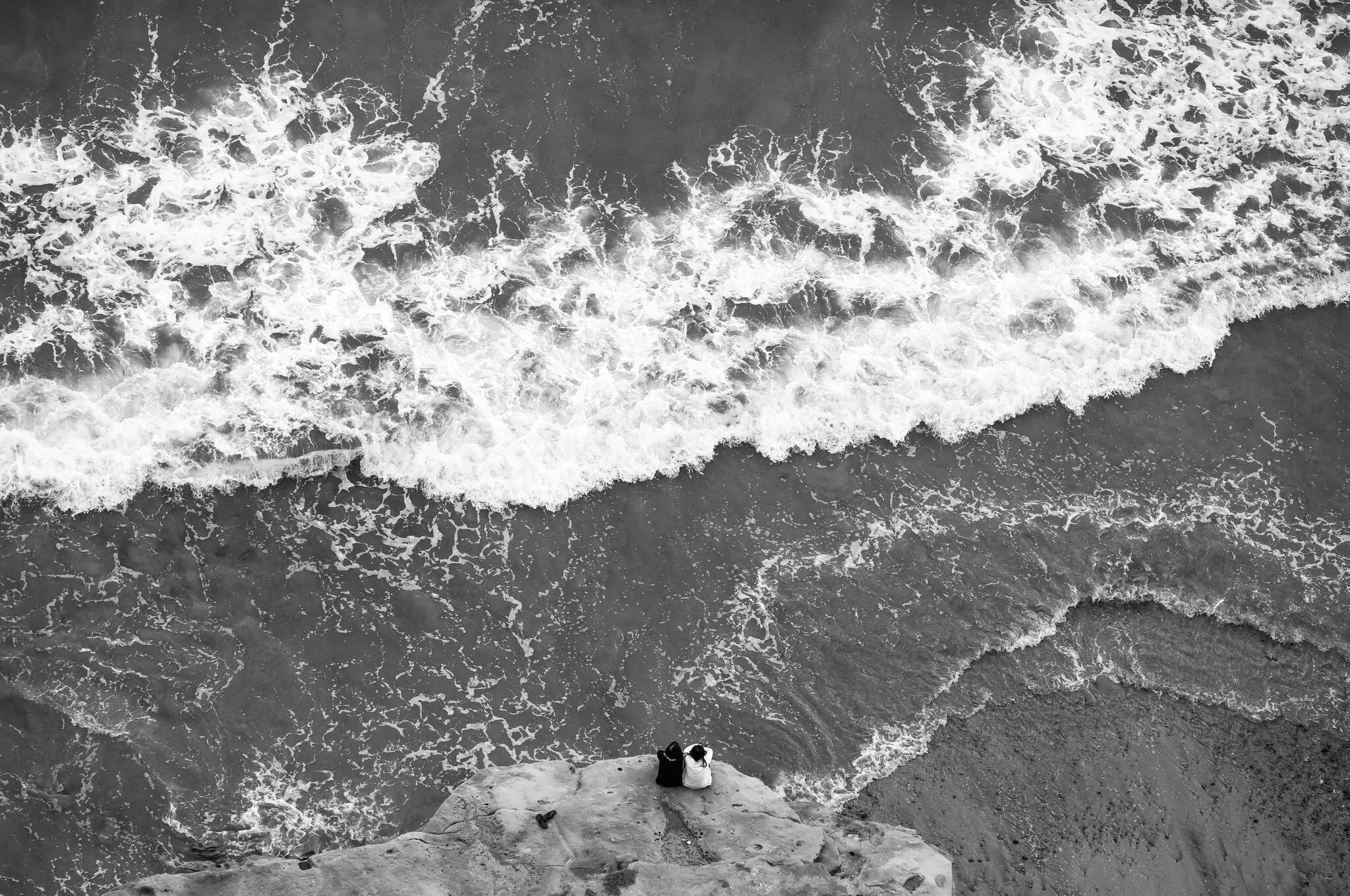 black-and-white photo of an aerial view of waves crashing into a beach and two people, sitting with their shoulders touching, gazing out into the waters
