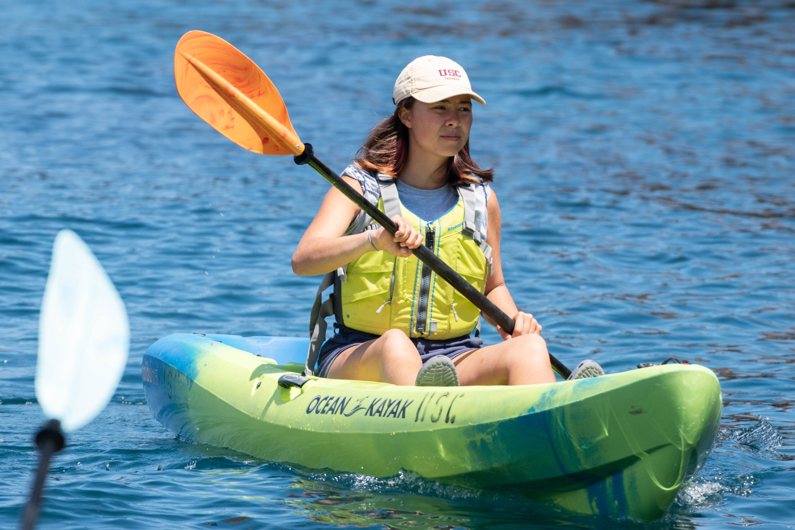 person wearing a bright yellow life vest and USC baseball cap sits on a kayak while paddling