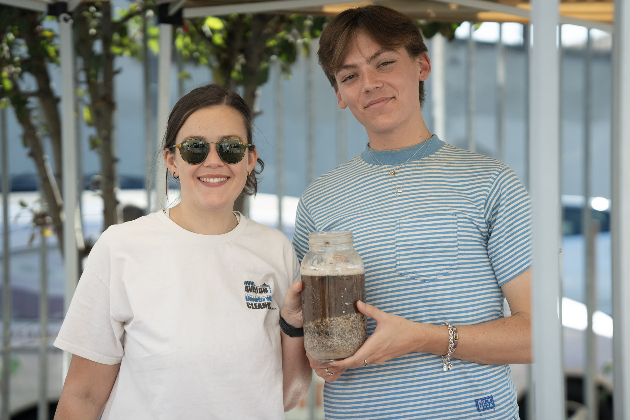 two people smile at the camera while holding a glass container of soil and rock