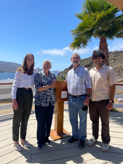 four smiling people stand on a wooden deck with ocean, hillsides, and a tree in the background