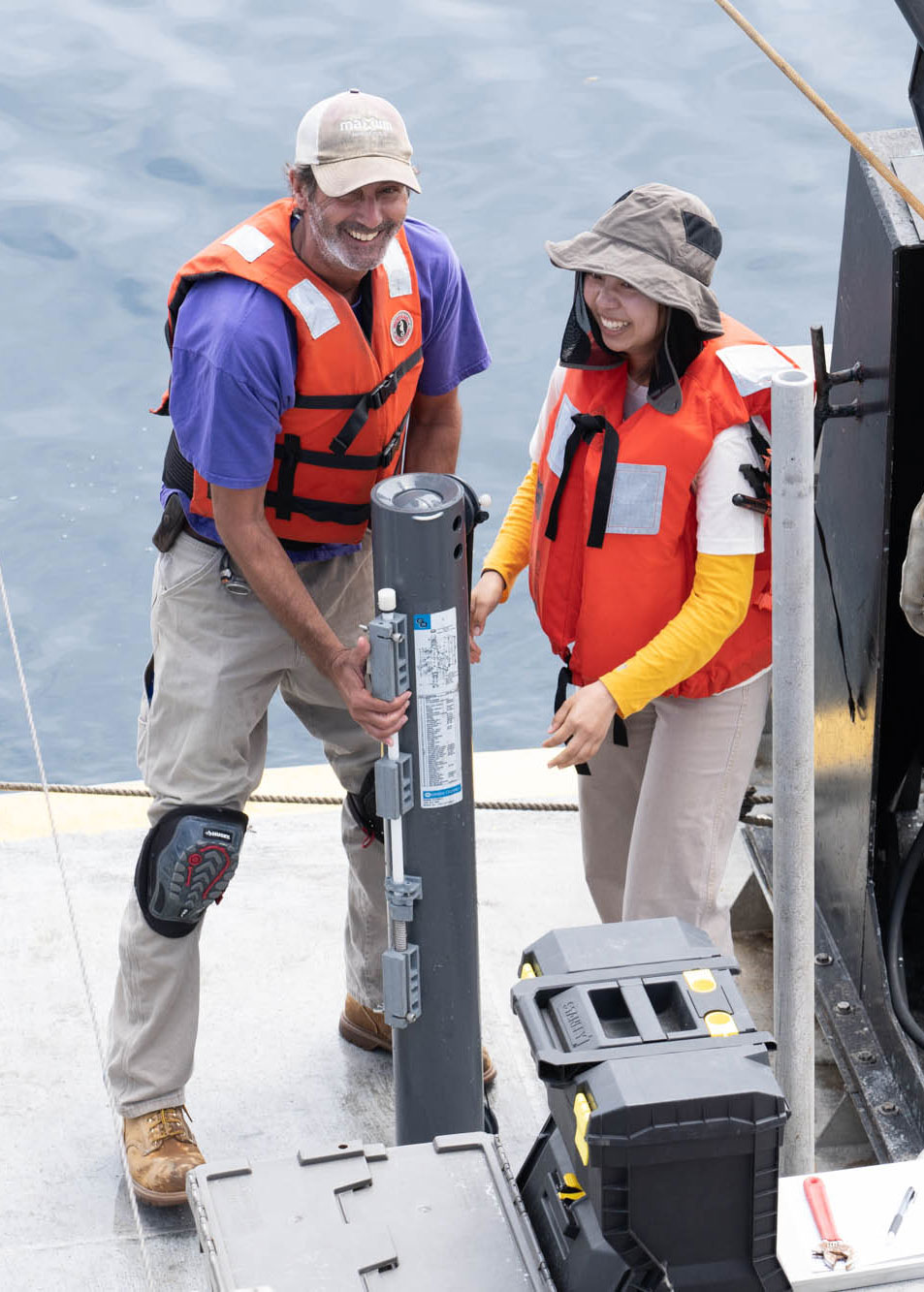 a smiling student and research assistant, both wearing bright orange vests, stand on either side of a dark gray cylindrical sampling tool on the deck of a research boat