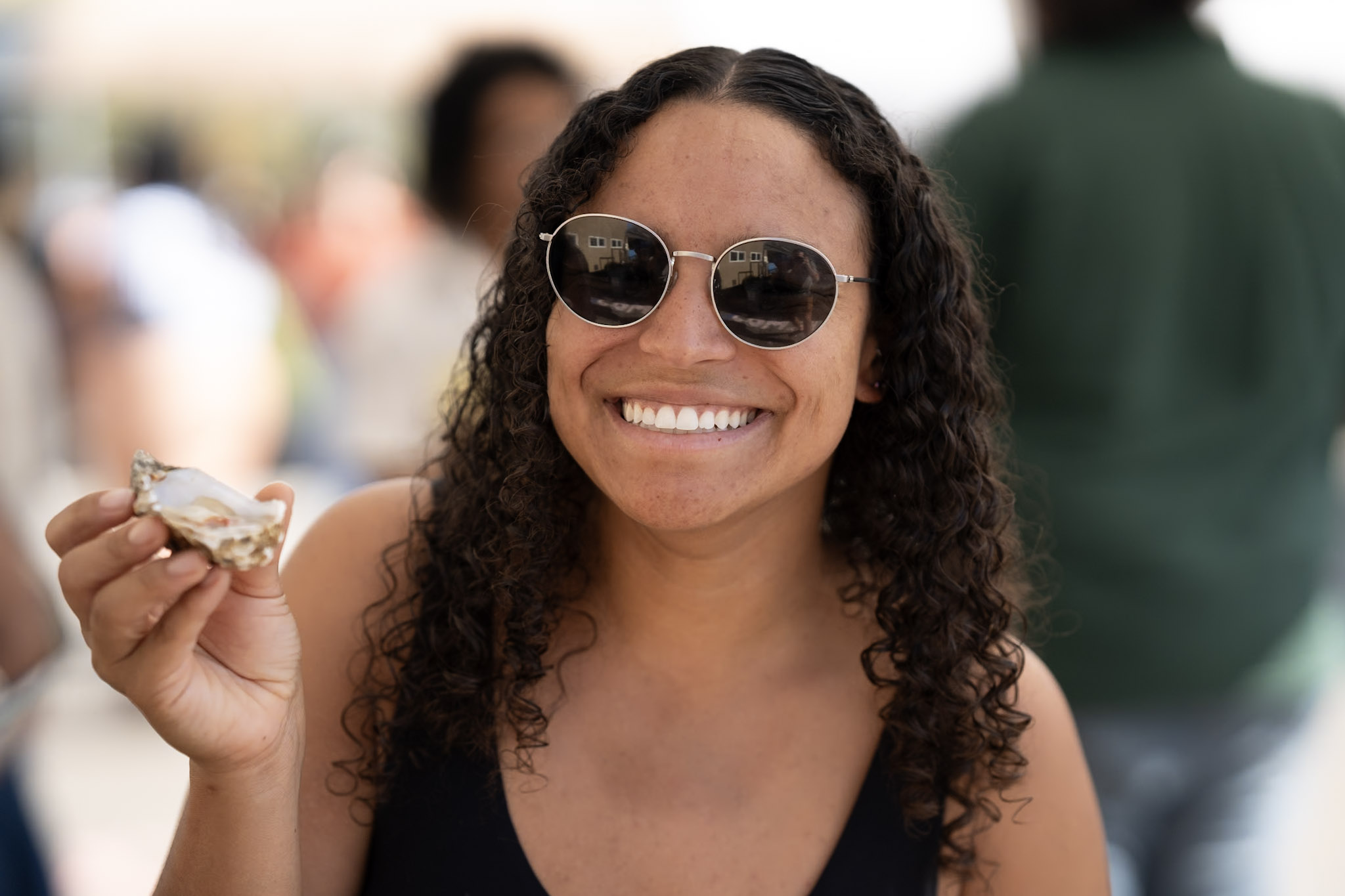 a person wearing sunglasses smiles at the camera while holding an oyster in one hand