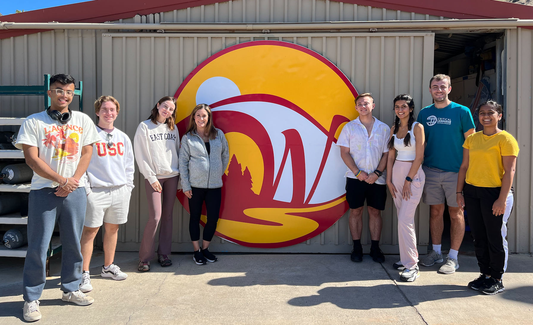 a professor and her students smile while standing in front of a large cardinal-and-gold "W" graphic on the side of a corrugated steel building