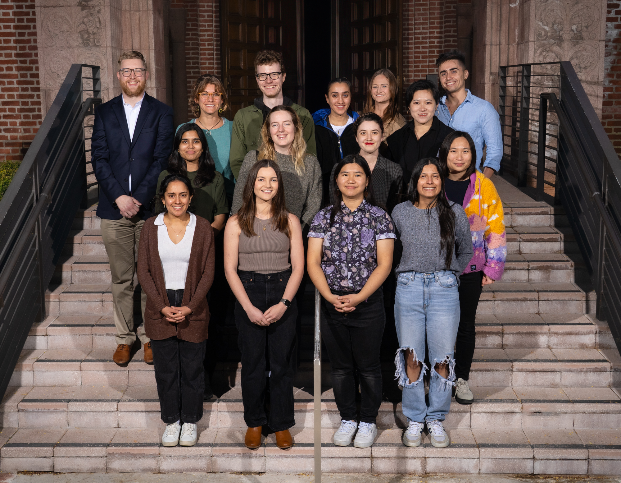 a diverse group of 15 smiling graduate students stand together on the concrete steps of a brick building