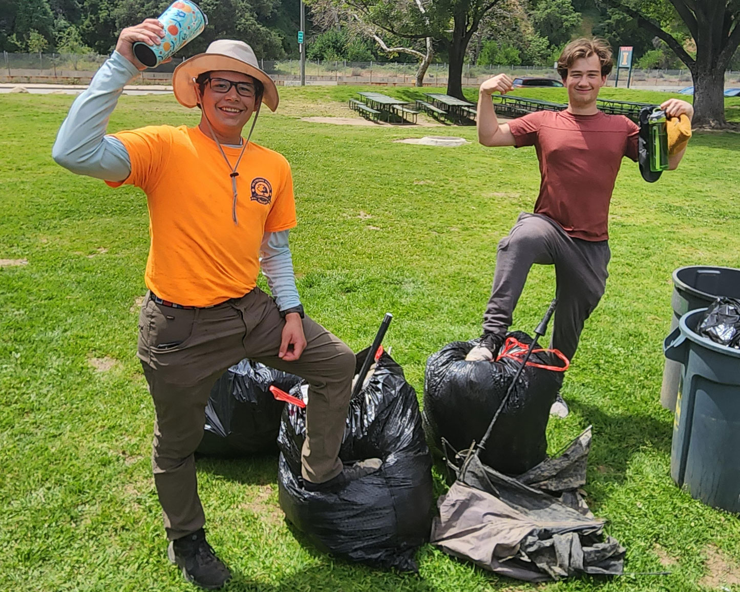 two students flex their arms and smile while posing with bags of plastic trash collected in a park