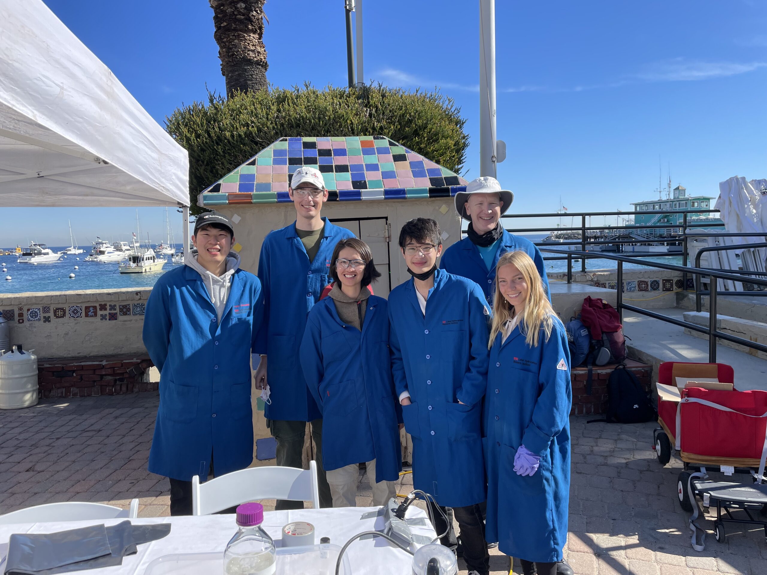 a professor and group of students wearing blue lab coats stand behind a table with an ocean harbor in the background
