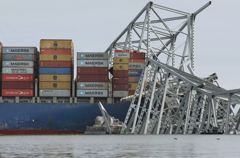 a blue cargo ship sits in the water, tangled in the steel wreckage of a large bridge
