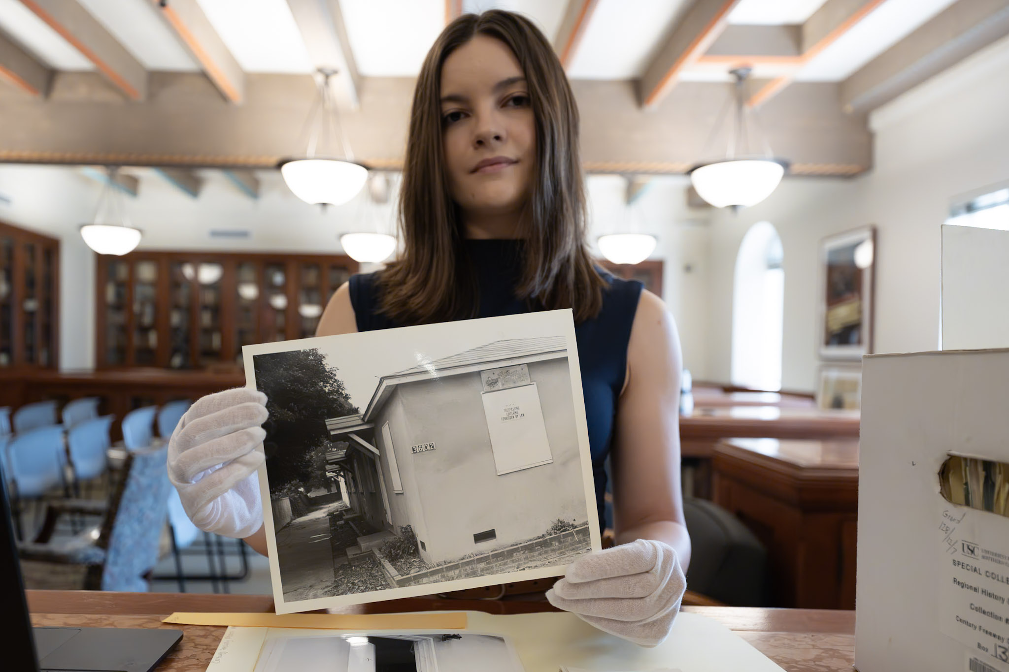 researcher wearing gloves holds a black-and-white photo of a house from the USC Libraries's archival boxes